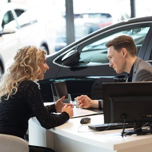 Consultants; A man and woman sit across from each other at a car dealership desk. They are engaged in discussion, with a parked car visible behind them.