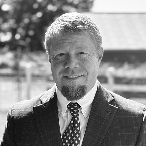 Cory Watson; A man in a suit and tie smiles warmly outdoors. The black-and-white photo has a blurred background of trees and a building, conveying a professional tone.