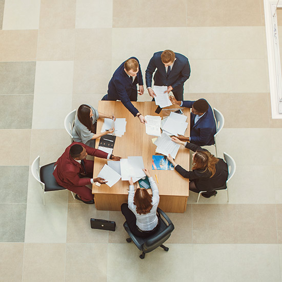 Staff; Aerial view of a diverse group of professionals collaborating at a rectangular table, exchanging documents, and engaged in discussion. Business setting.