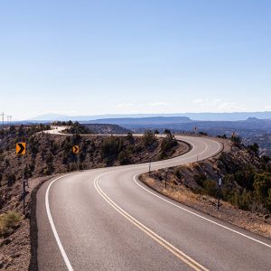 Utah Chapters; A winding mountain road curves through a barren landscape under a clear blue sky. Yellow arrow signs indicate sharp turns, creating a sense of adventure.