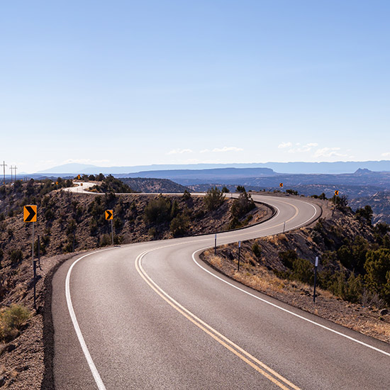 Utah Chapters; A winding mountain road curves through a barren landscape under a clear blue sky. Yellow arrow signs indicate sharp turns, creating a sense of adventure.